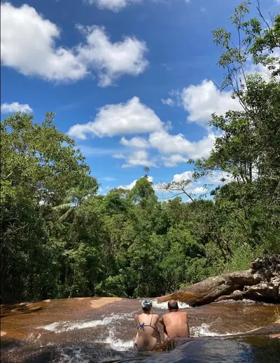 Casal sentado na beira de uma cachoeira contemplando a mata sob céu azul